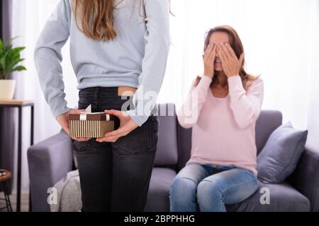 Girl Hiding Gift From Her Mother Sitting On Sofa And Covering Eyes Stock Photo