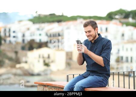 Happy man texting on smart phone sitting on a ledge on vacation in the coast Stock Photo