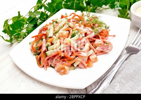 Salad from smoked sausage, spicy carrot, tomato, cucumber and spices, dressed with mayonnaise, napkin, fork and parsley on a light board background Stock Photo