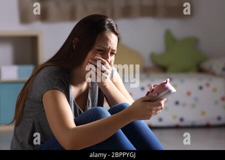 Sad mother missing her daughter holding a little shoe sitting on the floor of the bedroom in a house interior with a dark background Stock Photo