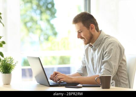Casual writer writing with a laptop at home sitting in a table near a window with a green background outdoor Stock Photo