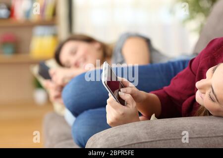 Two roommates using everyone their smart phones lying on a sofa in the living room at home with a homey background Stock Photo