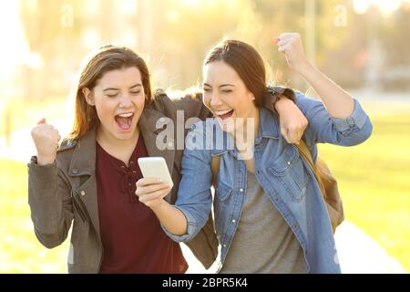 Two excited friends watching smart phone content in a park at sunset Stock Photo