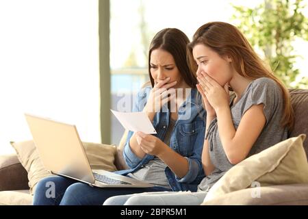 Two disgusted roommates reading a bank notification with bad news and checking on line sitting on a couch in the living room in a house interior Stock Photo