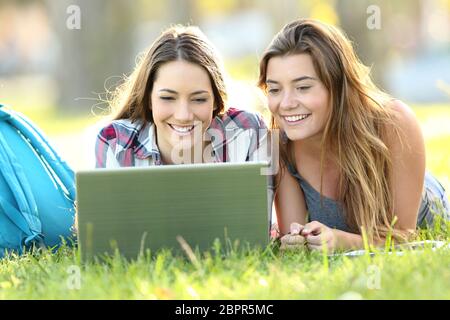 Front view of two happy students watching on line content with laptop lying on the grass in a park Stock Photo