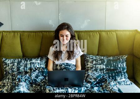 A smiling woman lying down the bed in front of her laptop with her legs raised slightly Stock Photo