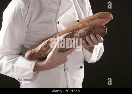 Baker in white uniform holding or carrying two baguettes of freshly baked rye bread, viewed in close-up against black background Stock Photo
