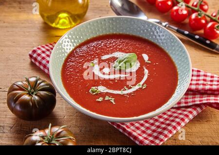 Plate of red tomato cream soup from high angle, served on red checked napkin with a spoon and different kinds of fresh tomatoes on rustic wooden table Stock Photo