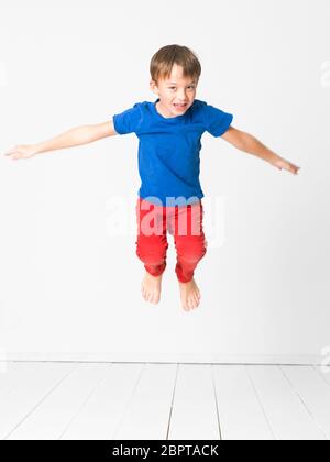 cool, cute boy with blue shirt and red trousers is jumping high in the studio in front of white background and white wooden floor Stock Photo