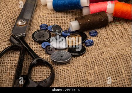 Several colored buttons of different sizes, reels of colored sewing thread, and large vintage sartorial scissors against a background of coarse-textur Stock Photo