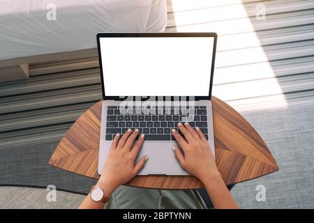 Top view mockup image of a woman using and typing on laptop with blank white desktop screen keyboard on wooden table next to the bed Stock Photo