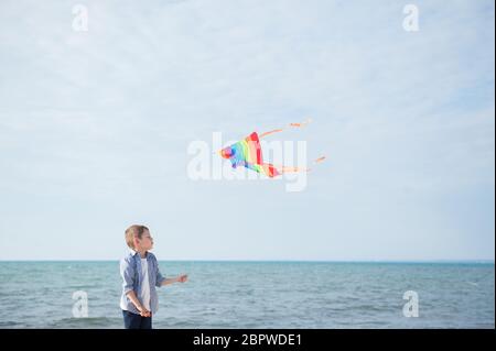 funny little boy with flying multicolored kite on blue sky and sea background with copy space Stock Photo