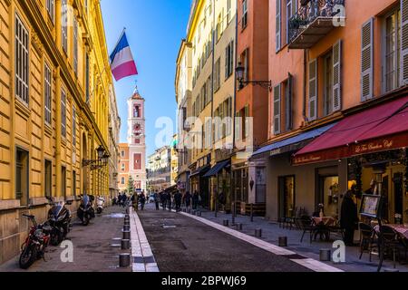 Nice, France, January 2020 – A typical pedestrian street of Nice old town with a French flag waving from a building and the Rusca Palace clock tower Stock Photo