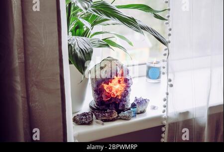 Using semi precious stone crystal details in home concept. Amethyst geode lamp burning, spiritual calming home atmosphere. Air plant Spathiphyllum. Stock Photo