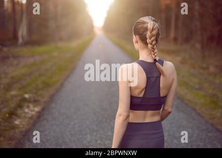 Sporty healthy young woman walking at sunrise along a rural road through a dense forest towards the glow of the sun at the end between the trees in a Stock Photo