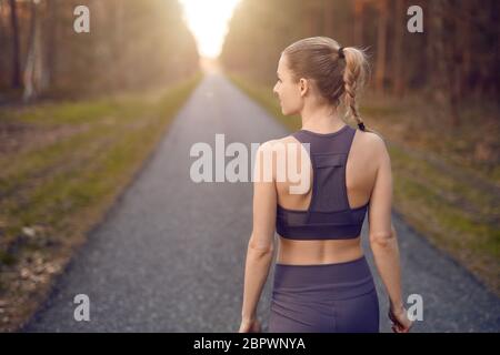 Sporty healthy young woman walking at sunrise along a rural road through a dense forest towards the glow of the sun at the end between the trees in a