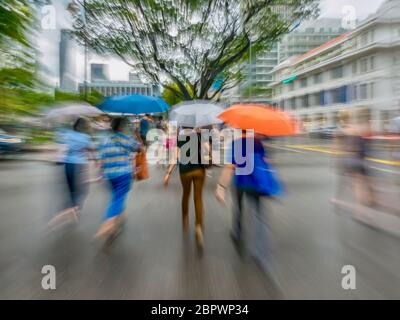 A motion blurred shot of pedestrians crossing a street on a rainy day in downtown Singapore. Fast paced city life. Stock Photo
