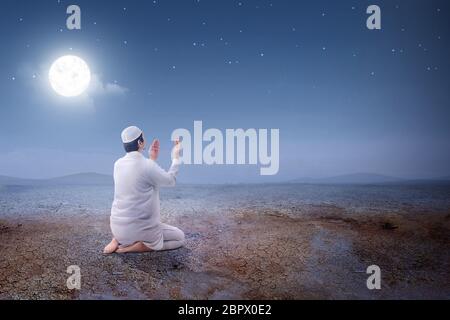 Rear view of asian muslim man sitting in pray position while raised hands and praying on the sand dune with moon and night scene background Stock Photo