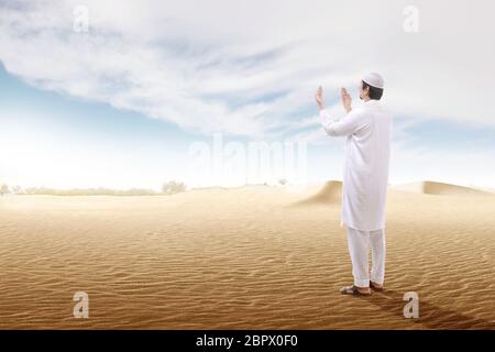 Rear view of asian muslim man raised hands and praying on the sand dune with blue sky background Stock Photo