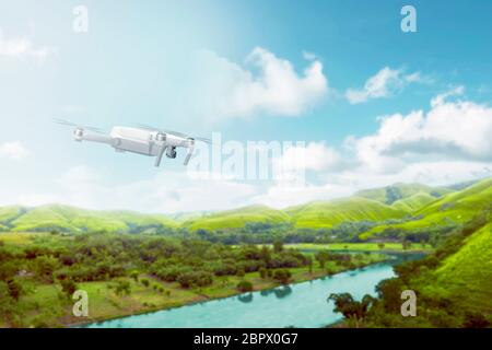 White drone with camera flying over green hills with river and trees over blue sky background Stock Photo