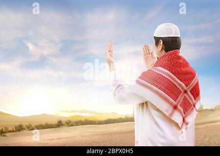 Rear view of asian muslim man with cap and turban standing and praying with raised arms on the sand with blue sky background Stock Photo