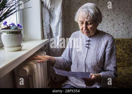 The senior woman holding gas bill in front of heating radiator. Payment for heating in winter. Stock Photo