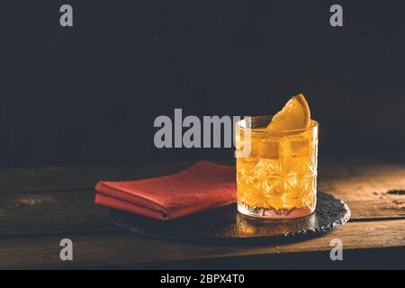 Glass of the cocktail negroni with red napkin and black stone tray on a old wooden board. Decorated orange slice. Nice romantic backlight. Stock Photo