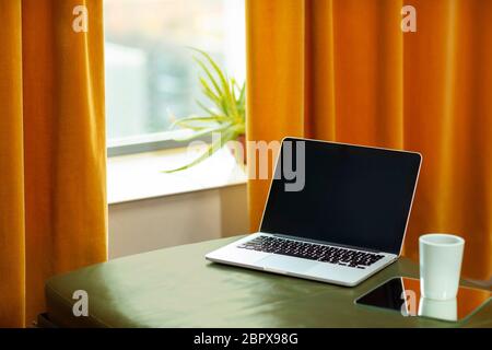 A laptop computer laying on a green couch and a orange curtain. A retro interior with warm colors and a cup of tea on the side. Modern technology and Stock Photo
