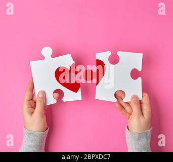 Female hands hold big white puzzles with a piece of red heart, pink background. Reunion concept Stock Photo