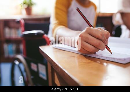Close up of female student taking notes sitting in wheelchair at desk. Focus on hand writing in notebook with a pencil. Stock Photo