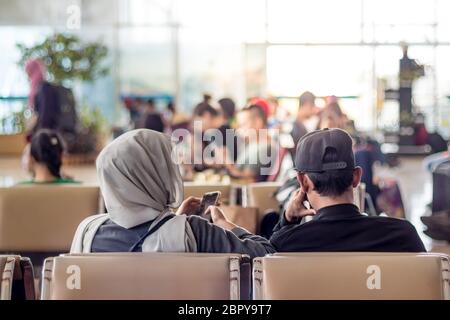 Modern muslim islamic asian couple using their smartphone apps while sitting and waiting for flight departure at international airport terminal. Stock Photo