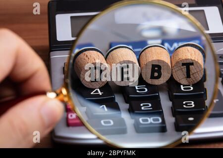Human Hand Examining Debt Word On Cork Through Magnifying Glass Over Calculator Stock Photo