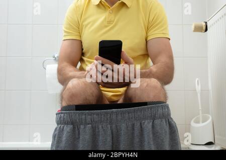 Mid-section Of Man Sitting On Toilet Using Smartphone Stock Photo