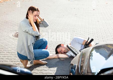 Young Woman Looking At Unconscious Male Cyclist Lying On Street After Accident Near Car Stock Photo
