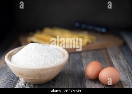 Bowl of whole wheat flour and two eggs on a rustic wooden table with pappardelle pasta in the background. Stock Photo