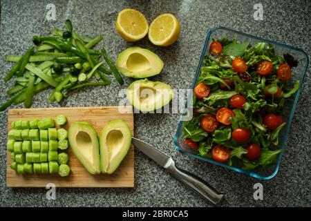 Preparation of a vegan salad with heart shaped avocado. Lemons, cucumbers, tomatoes, spinach, lettuces are on the table. There is also a sharp knife Stock Photo
