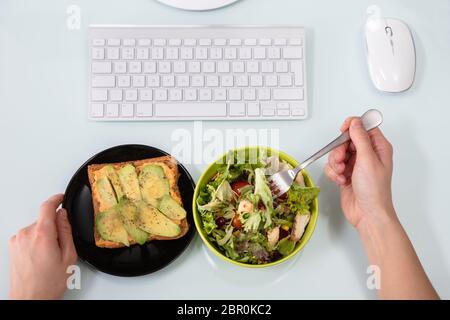 An Overhead View Of Businessperson's Hand Eating Salad With Fork On Desk Stock Photo