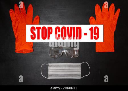 Black medical background. Safety goggles, medical mask and two red gloves. On the gloves there is a banner with the words Stop Covid-19 Stock Photo