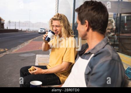 Two Caucasian male surfboard makers sitting on a bench and drinking coffee and having a snack Stock Photo