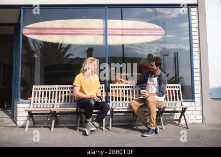 Two Caucasian male surfboard makers sitting on a bench and drinking coffee and having a snack Stock Photo