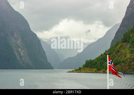 Flag of Norway waving in wind on a ferry that is heading away from land. Ship wake trace on water behind a vessel, distant mountains, beautiful evenin Stock Photo