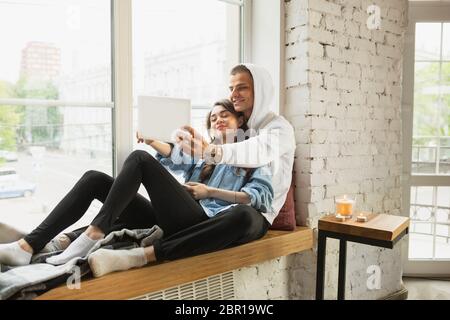 Hugging near window. Quarantine lockdown, stay home concept - young beautiful caucasian couple enjoying new lifestyle during coronavirus worldwide health emergency. Happiness, togetherness, healthcare. Stock Photo