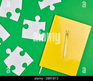 closed yellow notebook and pen, next to it are large empty white paper puzzles, green background, top view Stock Photo