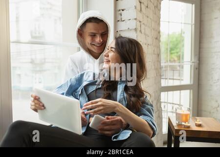 Hugging near window. Quarantine lockdown, stay home concept - young beautiful caucasian couple enjoying new lifestyle during coronavirus worldwide health emergency. Happiness, togetherness, healthcare. Stock Photo