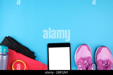 electronic tablet with empty white screen, next to it is fitness clothes, blue background, top view