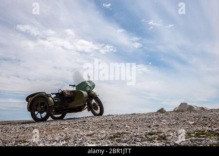 An old Soviet motorcycle with a sidecar stands against the sky with clouds on the stones. Green color. Windshield and round headlight. Horizontal. Stock Photo