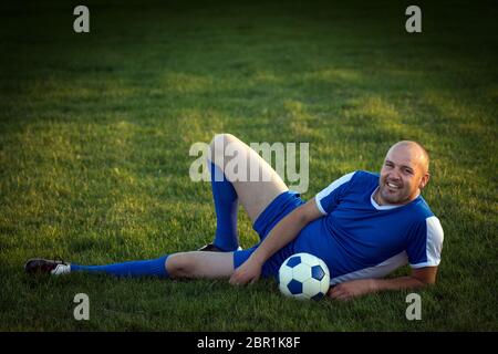 The smiling senior soccer player in blue dress is lying down on green grass on playing field with ball. Stock Photo