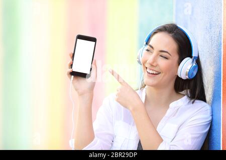 Happy girl listening to music showing and pointing smart phone blank screen in a colorful background Stock Photo