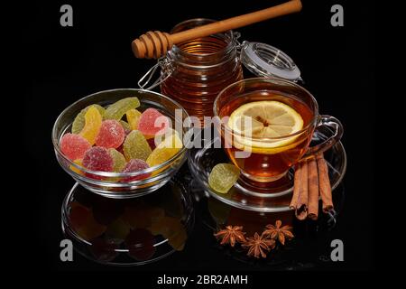 Honey in pot, fruit marmalade (fruit jelly candies) in glass bowl  and cup of tea with ginger, cinnamon, anise and lemon on a black background. Stock Photo