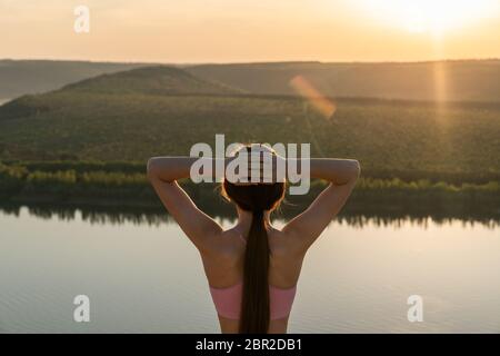 Waist up portrait of a beautiful sporty brunette female who stands back to the camera holds her hands on her head and enjoying sunset behinde the Stock Photo